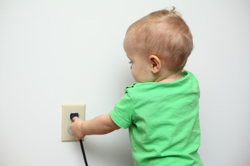 Electricity—How to Stay Safe. Baby in green shirt reaching to a pull a plugged in electrical cord in outlet.