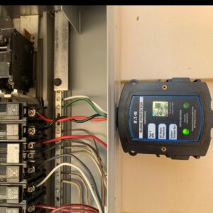 This image shows a whole-home surge protector installed adjacent to an electrical panel. The device, labeled with technical specifications, has indicator lights to display its operational status. The surge protector is connected to the panel with visible wires, which are neatly arranged, suggesting a professional installation.